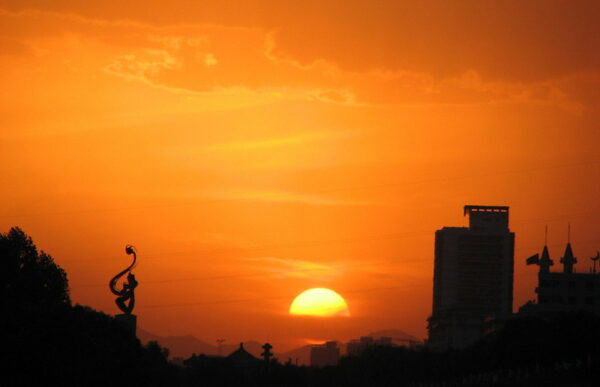 Against a vivid orange sunset and an enormous yellow setting sun wreathed with orange clouds, the trees and buildings of Lanzhou (the capital of Gansu Province, China) are but dark silhouettes.