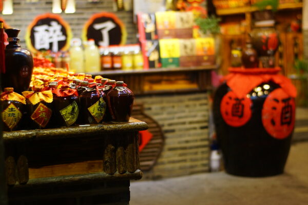 In a traditional Chinese-style shop, a wooden table on the left holds a display of small glazed pots of liquor, and on the right, there is an enormous glazed pot marked with red stickers indicating that it also contains some sort of liquor. The store is filled with other displays of boxes and bottles whose labels are not visible.