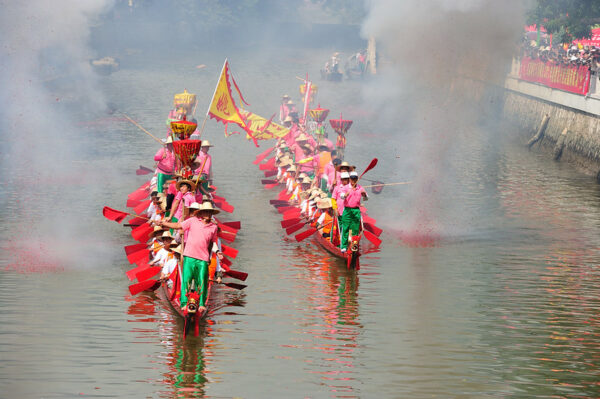 Smoke from firecrackers wreaths two festive red dragon boats, bedecked with red oars and red and yellow flags, as they sail side-by-side down a canal in Liede, Guangzhou’s annual Dragon Boat Festival. Each boat carries over 20 people, including standing crew members in bright green pants, pink shirts, and straw hats or baseball caps; and seated rowers wearing white shirts and orange life vests.
