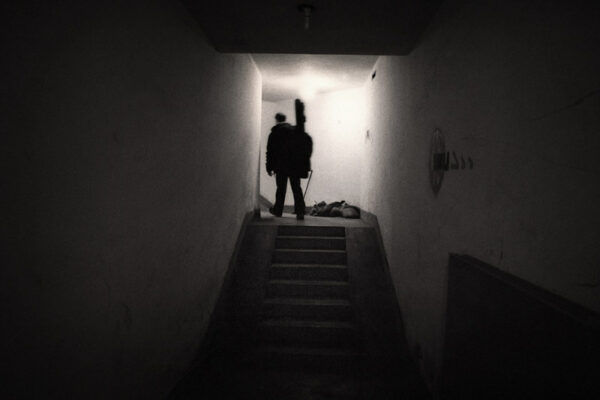 A grainy black and white image of a musician carrying a guitar case up a shadowy stairwell.