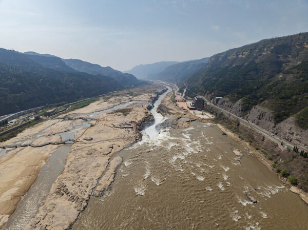 An aerial view of the Hukou Waterfall, the Yellow River’s largest waterfall. The wide and muddy river, flecked with large islands of yellowish soil, flows through a long river valley with low forested hills on either side. 