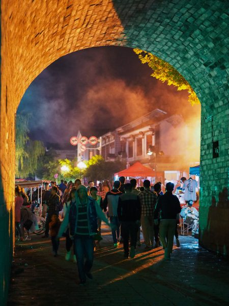 Evening scene of crowds passing through an arched tunnel that is one of the gates of the ancient city wall in the town of Dali, Yunnan Province. Beyond the tunnel are numerous stalls selling clothing and food; yellow and orange lights reflect off the smoke that fills the air.