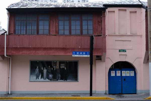 An old two-story pale pink building with a tin roof, a half-moon doorway with a bright blue metal gate, and a mural on its facade depicting nine historical figures, located along Shanghai’s Xinzha Road.