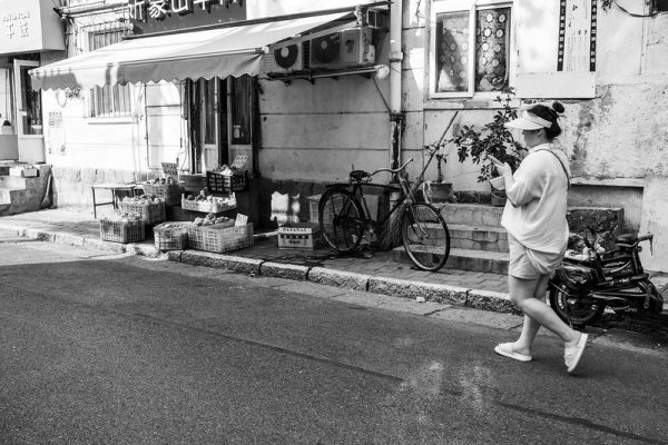 Black and white image of a woman dressed in light shorts, a t-shirt and a sun hat, texting while she walks down a narrow lane in Qingdao. Along the sidewalk are bicycles and a small shop displaying crates of fruits and vegetables.