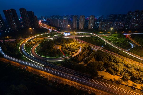 A long-exposure photo of a traffic interchange at night, surrounded by trees, with rows of tower blocks in the background