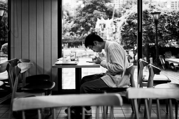 A young man wearing glasses, a white shirt, and dark slacks sits at a table in KFC, eating from a bowl of food as he hunches over a sheet of paper and a cell phone. On the opposite side of the table is a tray of KFC food.