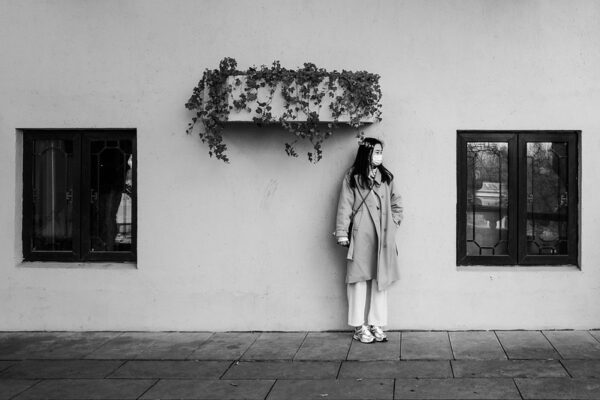 A young woman in a light-colored coat, trousers, tennis shoes, and a surgical mask stands against the wall of a building adorned with two windows and a planter filled with ivy. She stands off-center, much closer to the right-hand window.