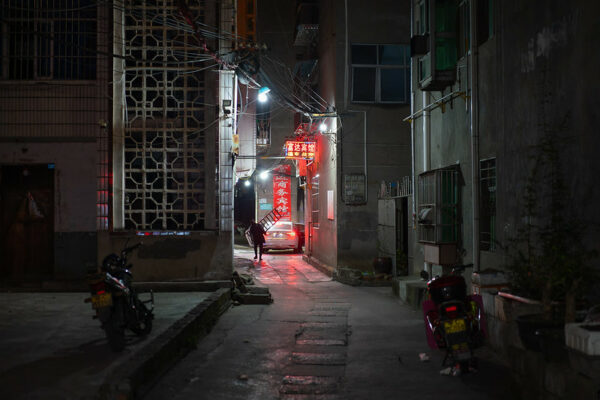 A narrow alleyway with residential buildings on both sides is lit by a red neon sign for a small guesthouse and the red tail lights of a car stopped at the end of the alley. In the glow of the tail lights, a man holding a ladder is visible, as are two parked mopeds in the foreground.