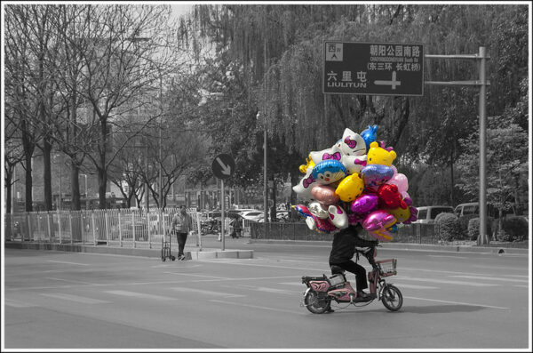 A man on a small electric bicycle crosses an intersection near Beijing’s Chaoyang Park with a large cloud of colorful helium mylar balloons shaped like well-known cartoon characters Hello Kitty, Doraemon, and Winnie the Pooh. The photo is black and white: only the bicycle, the bicyclist, and the balloons are in color.