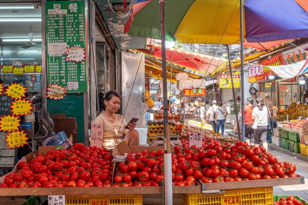 A woman dressed in shorts and a tan t-shirt looks at her cell phone while she sits behind a produce stall heaped with bright red tomatoes. Behind her, to the right, is a narrow alleyway lined with numerous other produce stalls, shop signs, and brightly colored canopies for shade. Photo taken at Baohua Avenue, Guangzhou, China.