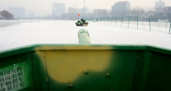 A view from inside a toy tank, whose “gun barrel” appears to be pointing directly at another toy tank that is being driven around a fenced-in field of ice and snow. A notice inside the tank, on the left-hand side, gives directions for turning left, turning right, braking, and driving. The sky is overcast and various multi-story buildings and a few trees are visible in the distance, through the haze.