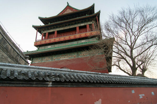 Behind a gray-tile-topped vermillion wall and a bare winter tree rises the elegant vermillion, green, and gold square-walled structure of Beijing’s famed Drum Tower.