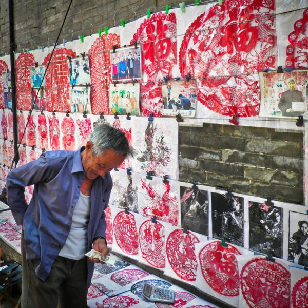 Standing in front of a brick wall hung with an array of intricate and finely crafted red paper cuts for sale, a gray-haired man in a blue shirt, white undershirt, and corduroy slacks holds a bill in one hand and reaches into his back pocket with the other hand. A calculator is visible on the ledge behind him.