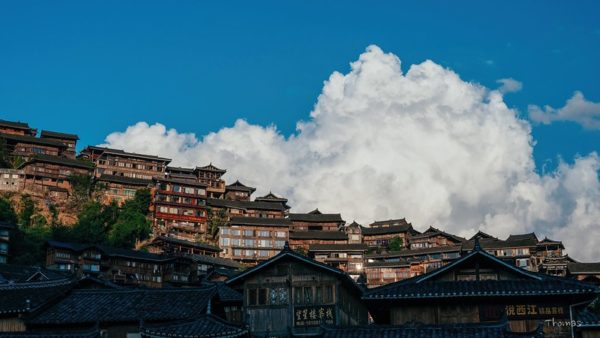 Beneath blue skies and white billowy clouds, a hillside is crowded with multi-story buildings in the traditional Guizhou style: wooden facades, tiled rooftops, ample windows, and intricate latticework.