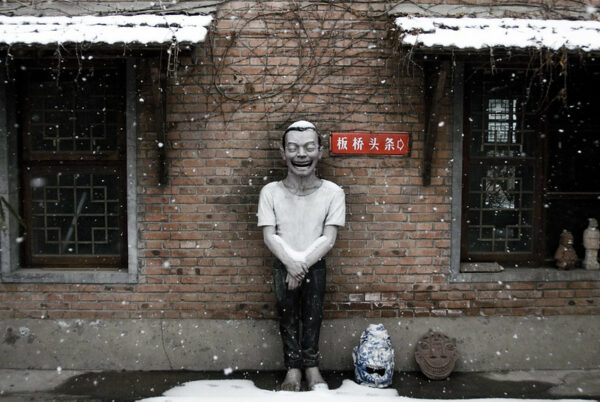 Falling snow accumulates on the head of a sculpture of a laughing man— with an enormous mouth and a large number of small and very even teeth—that has been positioned against the brick wall of a traditional Chinese courtyard home.