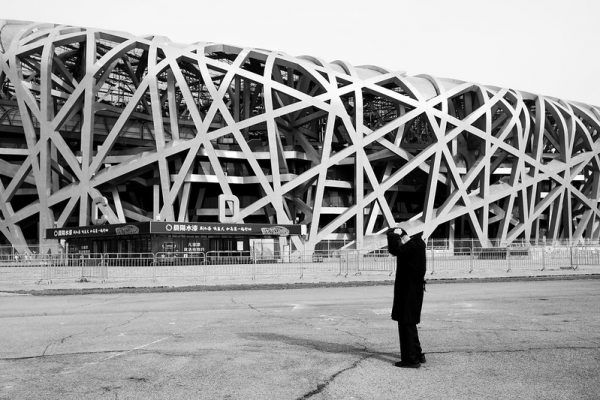A man dressed in black places a hand on the back of his bowed head as he stands before the tangled steel girders of Beijing Olympic Park’s National Stadium, known colloquially as the “Bird’s Nest.”