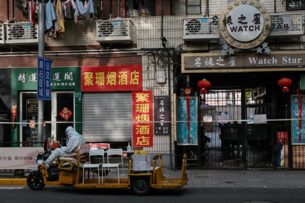 A long yellow motorized cart holding several white chairs is parked alongside an otherwise empty commercial street in Shanghai. The white-suited driver of the cart wears full PPE; the closed street is marked by a long tape strung between utility poles.