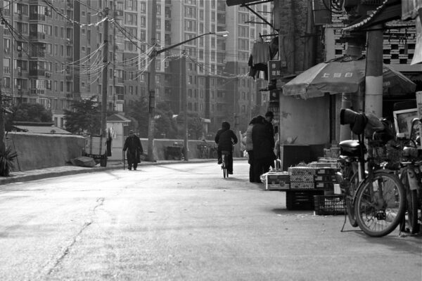 A bicyclist and several pedestrians pass along a curved street with neighborhood shops on the right hand side and tall residential buildings, streetlights, and power lines drooping from utility poles on the left hand side.