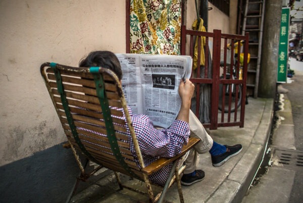 A man dressed in a blue and white checked shirt, khaki slacks, blue socks, and a pair of black tennis shoes crosses his legs casually as he pursues a newspaper while relaxing in a wood and metal folding chair on a narrow sidewalk along a Shanghai alleyway.
