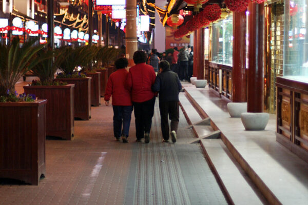 Three older women—two wearing red puffy jackets and one dressed in black—walk arm in arm through a shopping mall designed in traditional Chinese style and festooned with red lanterns and bright lights.