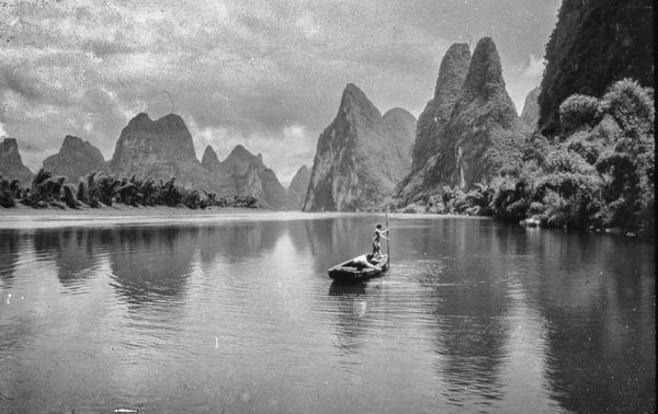 Under a clouded sky, man uses a long pole to pilot a small wooden skiff down the Li River, framed by the dramatic towering pinnacles of Guangxi’s famed karst hills.