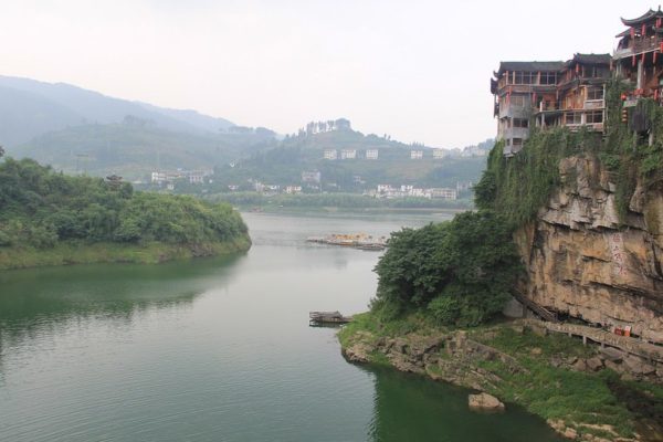 Traditional Chinese tile-roofed buildings perch atop high cliffs over a dark green body of water in the ancient town of Furong, Hunan province, known for its verdant hills and abundant waterfalls.
