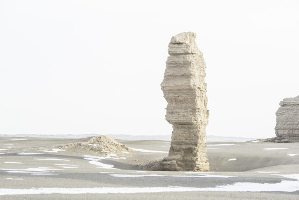 Against a whitish sky, a pale sandstone pillar towers like a monolith above the desert sands.