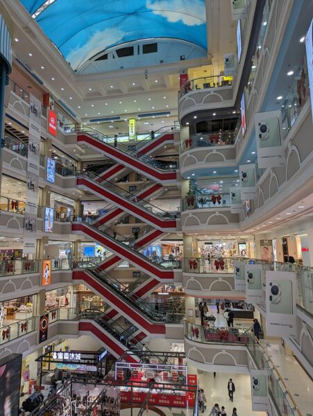 Red and white escalators crisscross the atrium of a gleaming new six-story shopping mall with five stories of shops and a basement level selling food and clothes. At the very top, an arched ceiling is adorned with canopies painted with blue skies and white clouds.
