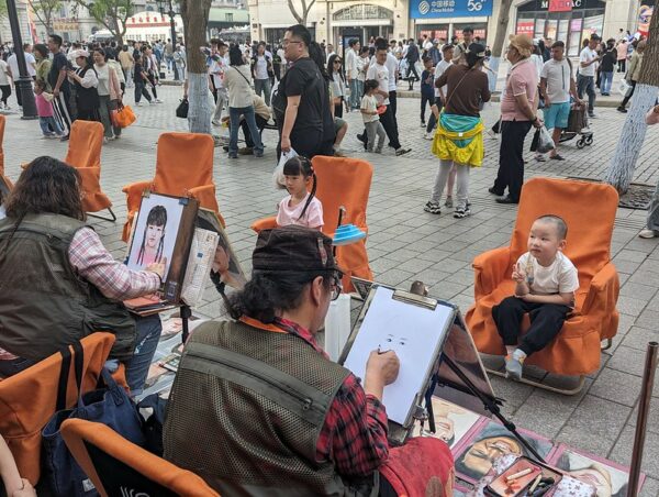 Along a busy shopping street in Harbin lined with stores and thronged with shoppers and pedestrians, two streetside artists draw portraits of two children—a little girl with braids and a little boy with a closely-cropped haircut—who are sitting in folding chairs covered with orange cloth chair covers.