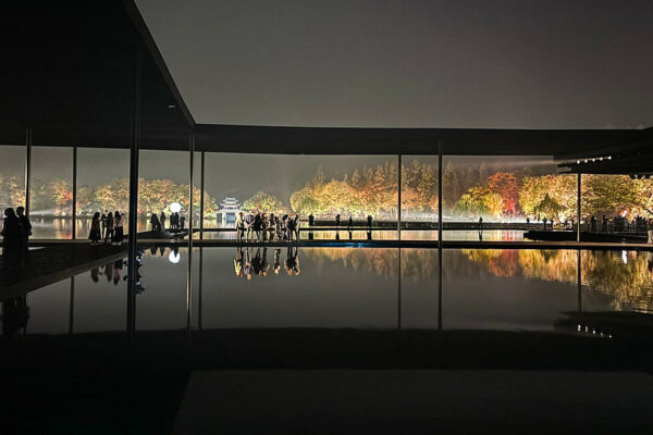 A stylized photo of a modern structure in the foreground, composed of catwalk-like walkways that seem to float over a body of water. Several groups of people standing on the catwalks appear to be reflected in the water below them. In the background is an illuminated vista of a traditional Chinese garden at night, with a half-moon shaped bridge and pagoda-like structure at the center, and a profusion of trees in shades of red, yellow, and green.