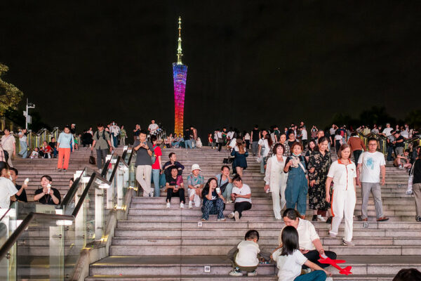 A long flight of concrete stairs in Guangzhou’s Huacheng Square is crowded with people walking, sitting, eating, drinking, chatting, and taking photos. At the top of the stairs, in the distance, the towering silhouette of Canton Tower is illuminated with festive, rainbow-hued lights.
