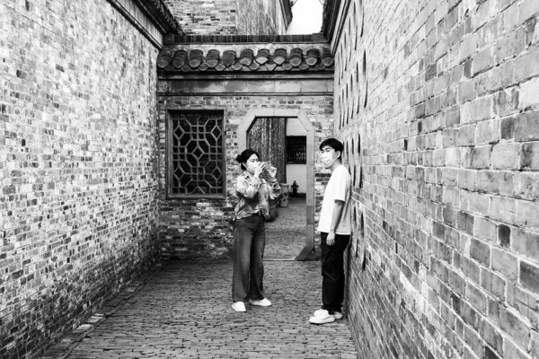 A young woman aims her cell phone camera at a young man leaning against the brick wall of a narrow cobbled corridor of a traditional Chinese one-story home. Both of them are wearing masks.