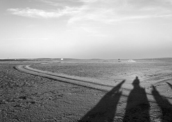 Long, oddly shaped shadows fall across a dirt road that winds across a mostly featureless landscape, somewhat resembling a moonscape.