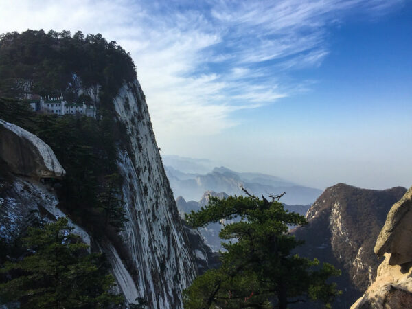 A stunning view of a monastery perched on the edge of a sheer cliff, dramatic pine trees, and verdant hills shading off into the distance.