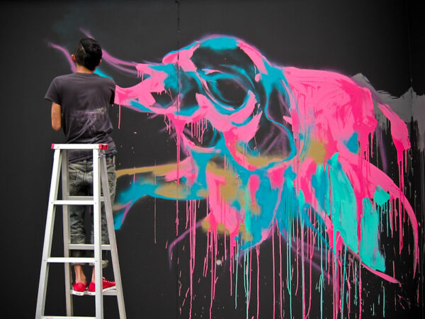 A short-haired artist in red tennis shoes standing on a ladder uses neon pink, blue, and turquoise spray paint to create a surreal elephant-like image on a black wall.