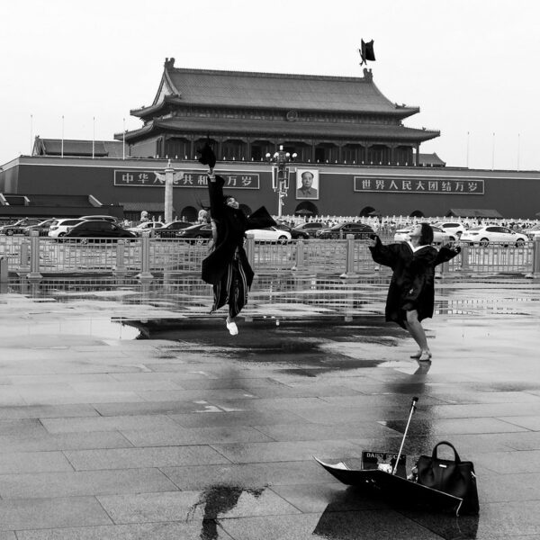 Two young women, dressed in dark-colored academic robes, joyously toss their graduation caps into the air on a rainy day in Beijing’s Tiananmen Square. Forming the backdrop is Chang’an Avenue, crowded with passing cars, and behind that, the iconic Tiananmen Gate with its portrait of Mao Zedong at the center.