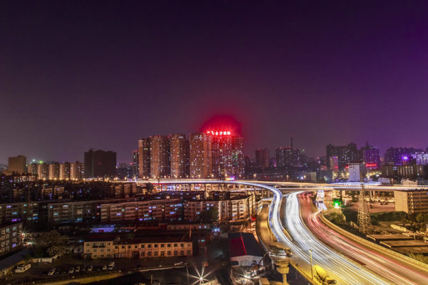 Long exposure shot of Zhengzhou’s cityscape at night features a deep purplish sky, winding expressways, and tall residential buildings with enormous red neon signs glowing atop them.
