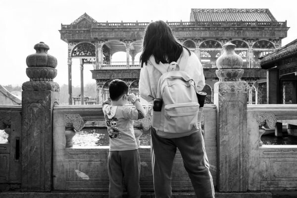 A woman with a backpack and a young boy with his fingers in his ears stand on a stone bridge, gazing out at an elegant, two-story, traditional Chinese pavilion across the water. The pavilion features elaborate carvings, pillars, arches, and a tile rooftop.