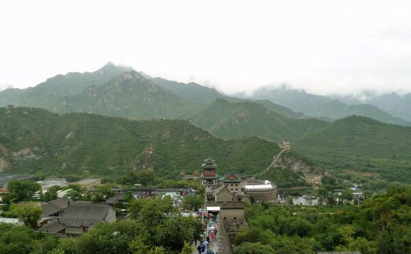 An aerial panorama of the Juyong Pass reveals tiny figures with umbrellas walking along the wall, and a collection of charming tile-roofed buildings of gray brick and red lacquer nestled among the verdant hills and low cloud cover.