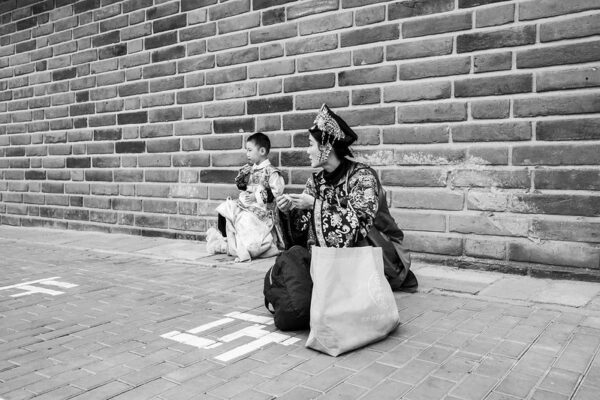 At Beijing’s Forbidden City, a woman and a young boy, both dressed in elaborate historical Chinese costumes, squat on the ground, as if taking a break. The woman has a modern-style backpack and canvas tote bag on the ground in front of her, which clashes somewhat with her silk brocade costume and beaded headdress.
