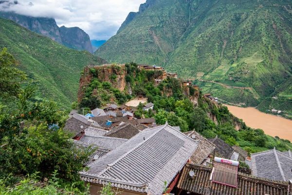 A bird’s eye view of the tile-roofed houses of Baoshan Stone Village in Yunnan Province, southwestern China. The houses descend along the ridge of a mountain; in the distance are more verdant mountains, a muddy river, low clouds and a spot of blue sky.