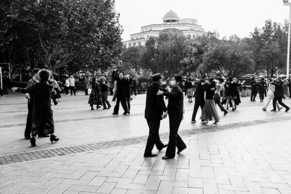 Several dozen middle-aged and older couples, some wearing surgical masks, dance during the daytime in a tree-lined public square in Qingdao.