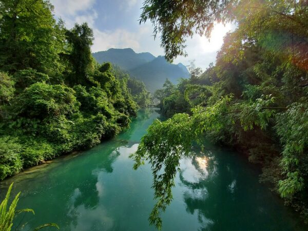 An emerald-colored river with lush vegetation on both banks wends into the distance, with Seven Arcs Bridge faintly visible, and beyond that, a series of deep green hills. The bridge is located in Libo County, Guizhou province.