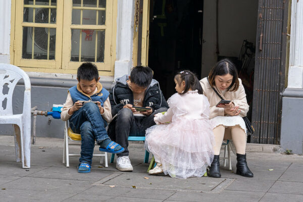 Seated on low folding chairs, two boys in jeans and puffa jackets and a woman in a white dress and black boots stare at their cell phones, immersed in what they are looking at on their screens. A younger girl wearing pigtails, tennis shoes, and a fancy white dress, but without a phone, looks at the boys sitting next to her.