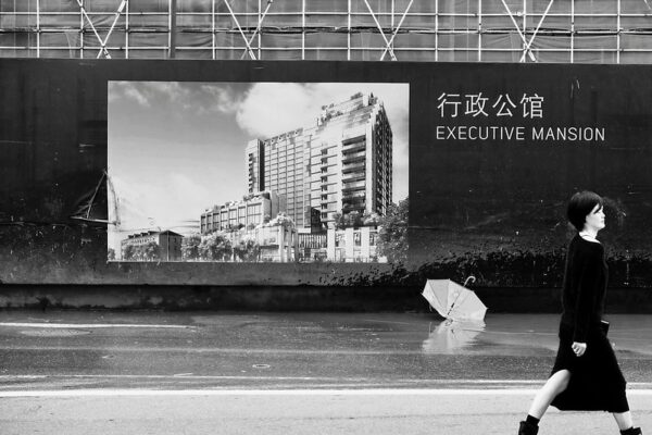 A woman with stylish short hair, a black dress, and black boots walks past a construction site and its advertisement, which shows an artist’s rendering of a modern highrise building with the tagline “Executive Mansion.” An upended white umbrella, seemingly abandoned, rests on the sidewalk below the advertisement.