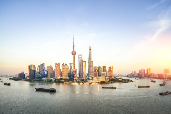 Sunlight reflects from what appears to be an island of the skyscrapers on The Bund in Shanghai, as enormous cargo ships navigate along the river surrounding it.