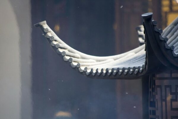 In this close-up, smoke and bits of ash drift upwards through the air toward one corner of a “winged” tiled rooftop at a Shanghai temple.
