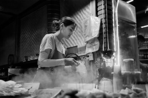 A ponytailed street food vendor, surrounded by steam from cooking, places some food in a plastic takeaway bag.