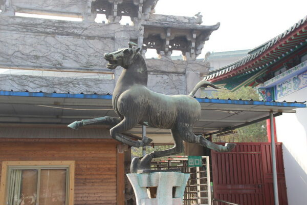 A whimsical bronze sculpture of a galloping horse is on display in what appears to be the courtyard of a traditional tile-roofed building.