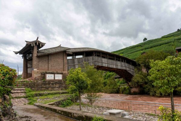 A wooden bridge constructed of dark ash-colored slats and a tiled roof arcs across a narrow, turbid river and disappears into the abundant bushes of a green hillside on the other side of the river. The nearest entrance to the bridge, where it meets the shore, is constructed of red brick and features a more elaborate tiled rooftop. There is also a stone retaining wall at the bottom of the brick entrance, and a series of irregular stone steps leading to a narrow path that winds along the riverside.